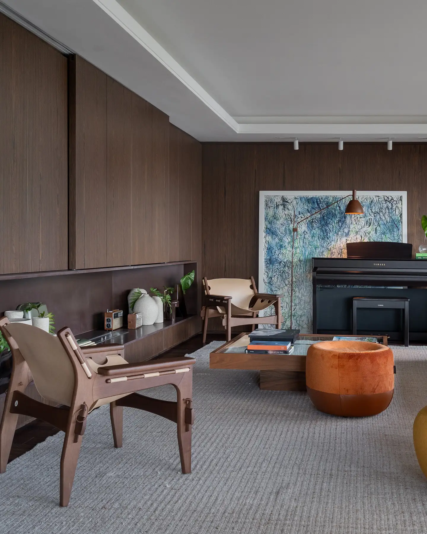 Living room with dark wood panel walls, mid-century chairs, and a black Yamaha piano against an abstract blue artwork backdrop under track lighting.