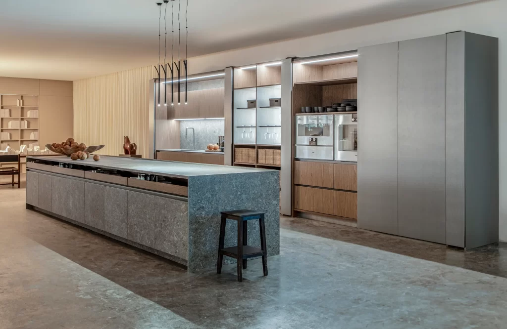 Modern open-concept kitchen showroom with a large granite island, wooden cabinets, and built-in appliances along the wall.